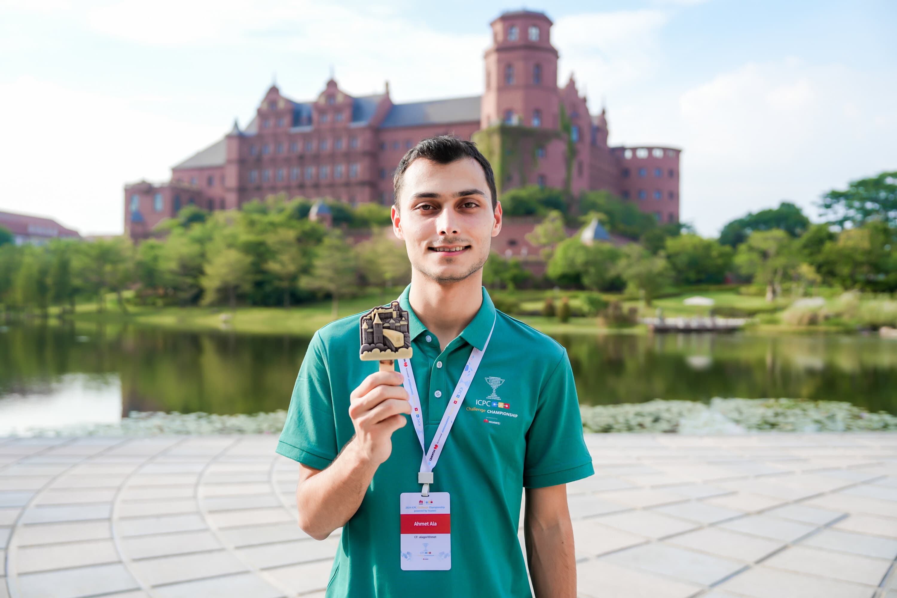 Ahmet Ala at the 2024 ICPC Challenge Championship in Shenzhen, holding an ice cream shaped like the Polybahn funicular, with the venue's campus behind him.